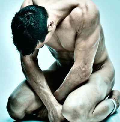 A hot man is seated on the ground and is showing off his nude body.